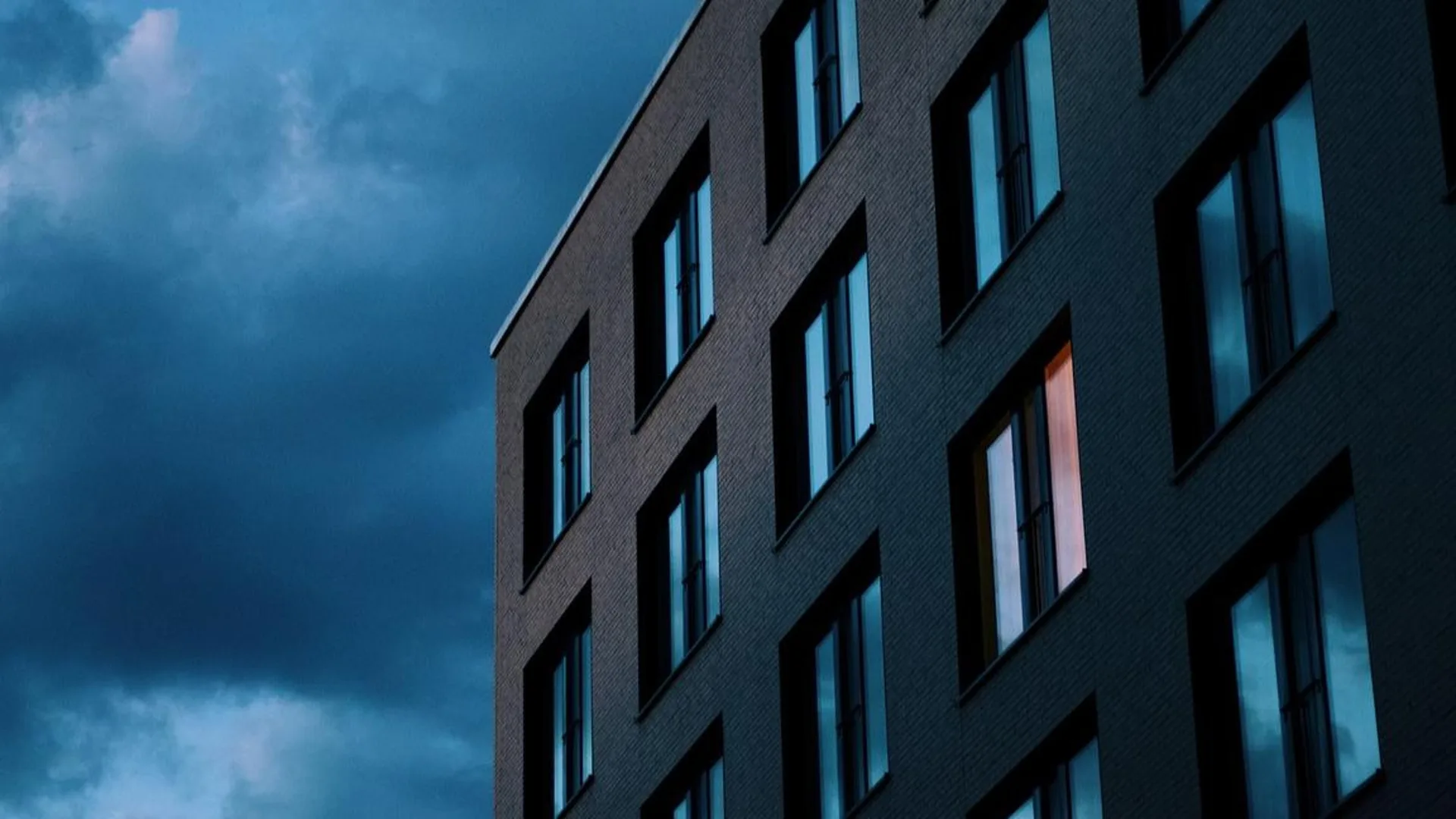 Moderner Wohnblock aus dunklem Backstein vor dämmerblauem Himmel, in einem Fenster brennt Licht