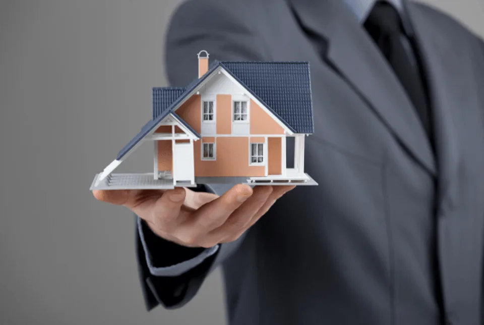 Ein Mann im Anzug hält ein kleines Modellhaus auf der flachen Hand